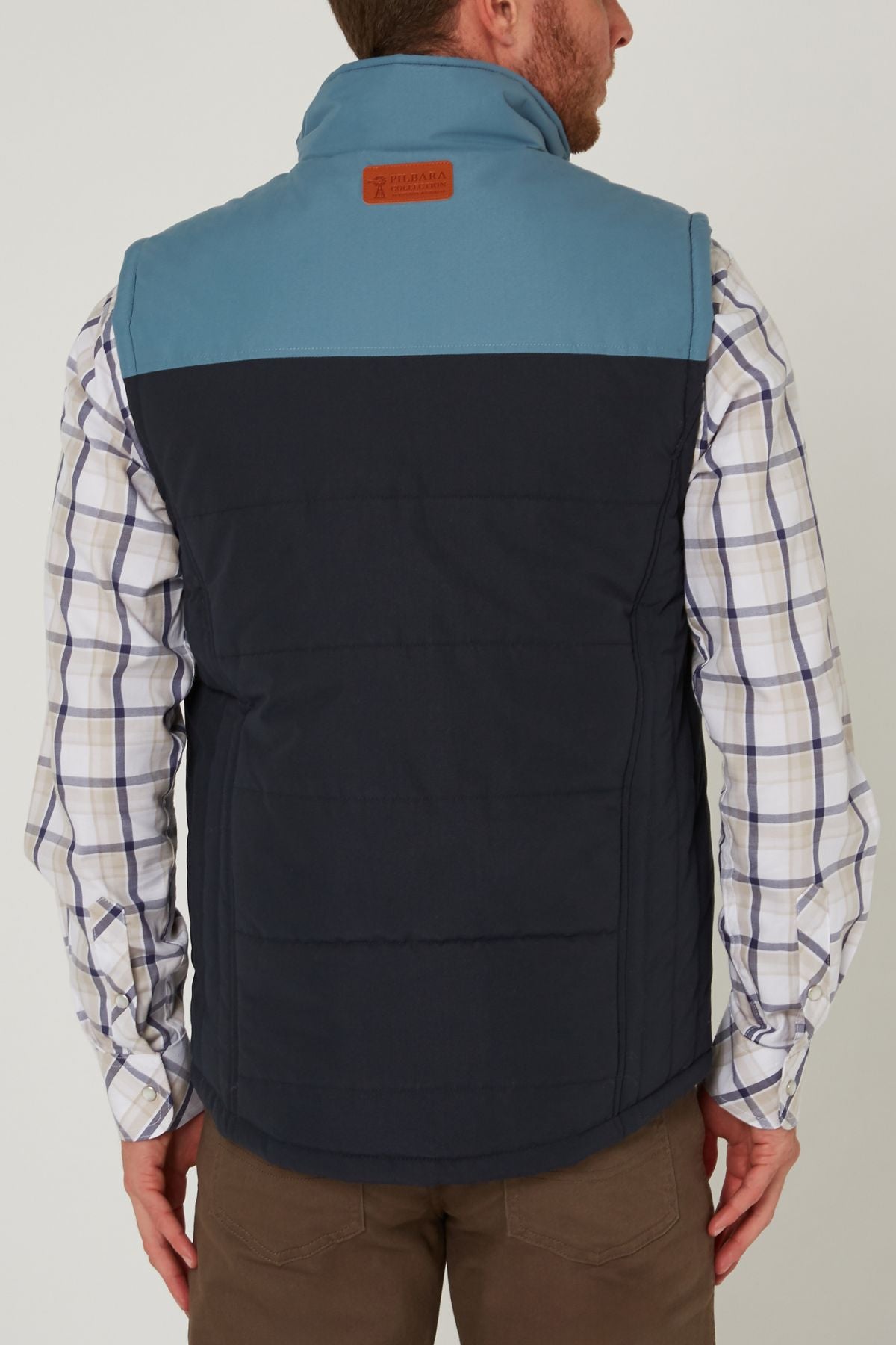A man shown from the back wears a Pilbara Two Tone Vest in blue and dark navy with polyfill padding over a plaid shirt and brown pants. The vest features a leather patch on the upper back.