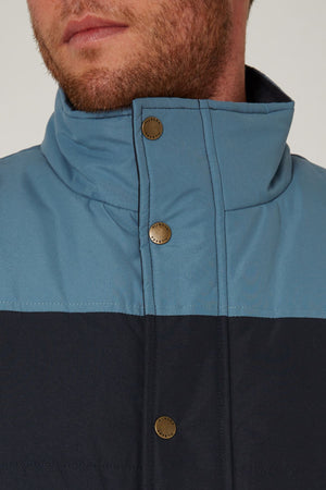 A close-up of a man's lower face and neck shows the Pilbara Two Tone Vest in blue and black, featuring metal snap buttons, polyfill padding, and a high collar. The man has light skin and a short beard.