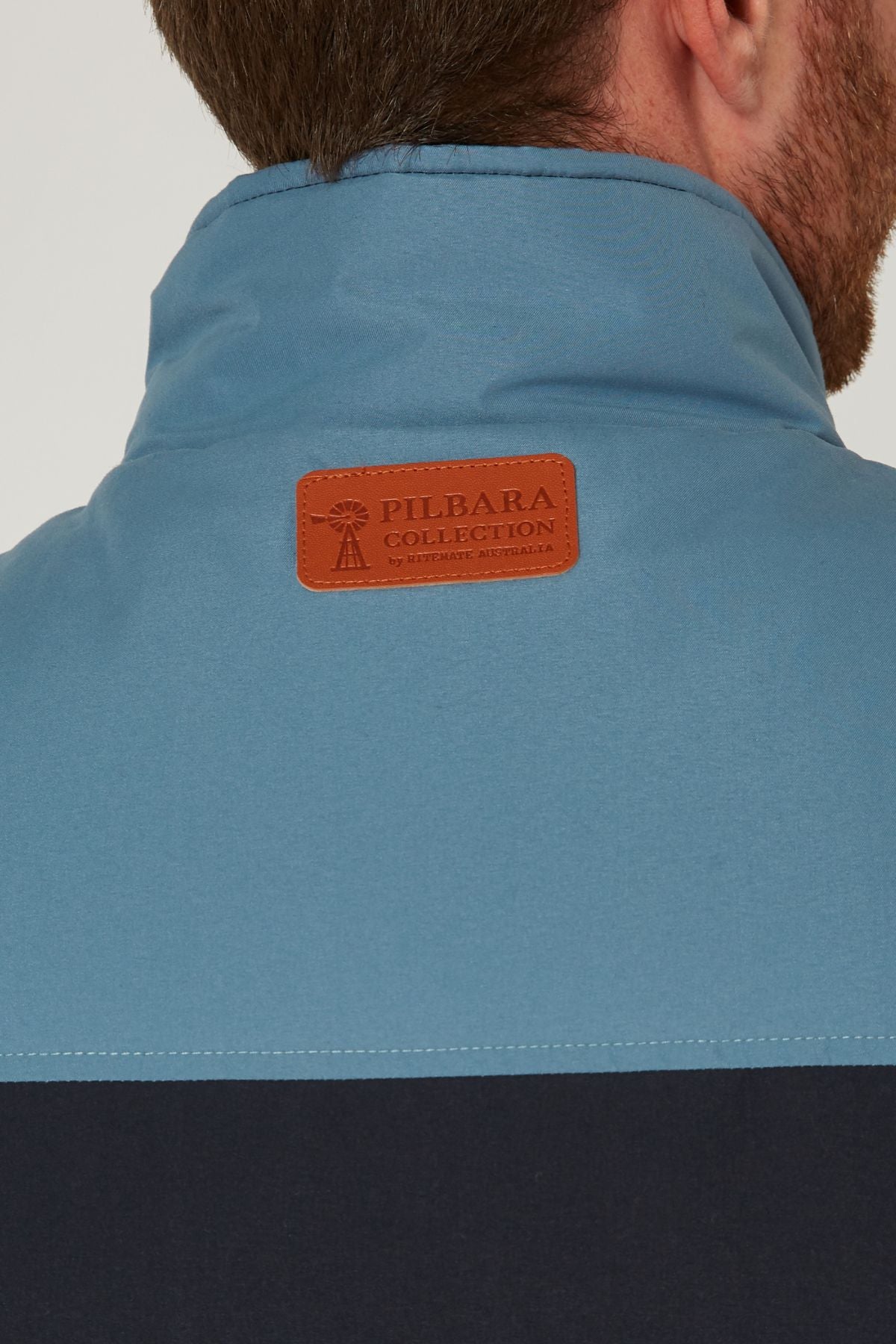 Close-up of the back of a person's neck and shoulders wearing a blue Pilbara Two Tone Vest. A brown leather patch with "PILBARA COLLECTION by Ritemate Australia" is sewn near the collar.