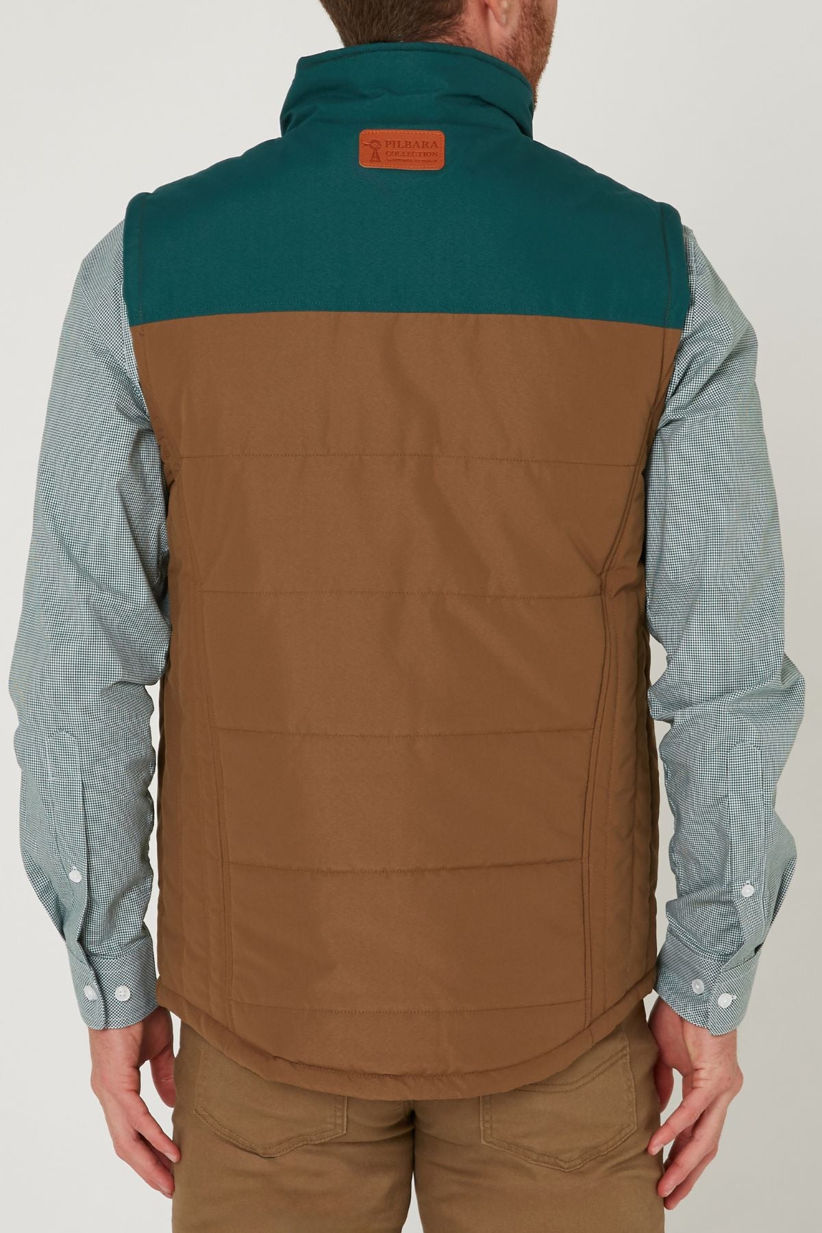 A man, seen from behind, wears the Pilbara Two Tone Vest in teal and brown over a light blue button-up shirt with khaki pants. The vest features a leather patch near the collar.