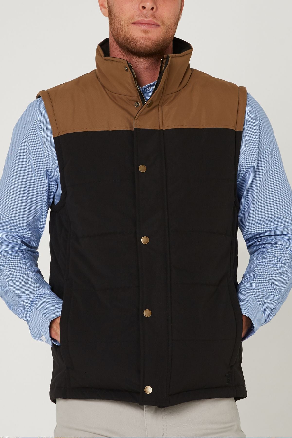 A man wears the Pilbara Two Tone Vest—a brown and black classic fit quilted vest with polyfill padding, stand-up collar, gold snap buttons, and pockets—over a light blue button-down shirt.