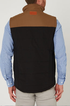 A man is seen from behind wearing the Pilbara Two Tone Vest, a classic fit padded brown and black vest with polyfill fill, over a light blue button-up shirt and beige pants. The vest features a brown patch on the upper back near the collar.