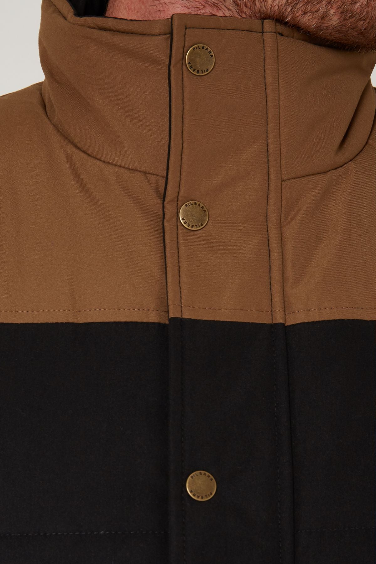 Close-up of a person's neck and upper chest wearing a high-collared Pilbara Two Tone Vest in brown and black, featuring three metallic snap buttons on the front.