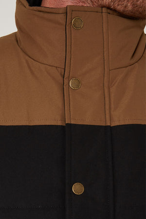Close-up of a person's neck and upper chest wearing a high-collared Pilbara Two Tone Vest in brown and black, featuring three metallic snap buttons on the front.