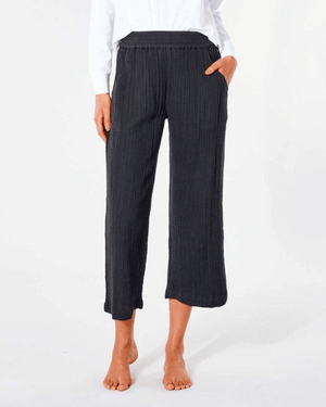 A person in a white long-sleeve shirt wears Rip Curl Premium Surf Beach Pants, black textured with a cotton double weave. These mid-calf pants have an elastic smocked waist. They're barefoot against a plain white background, hands in pockets.