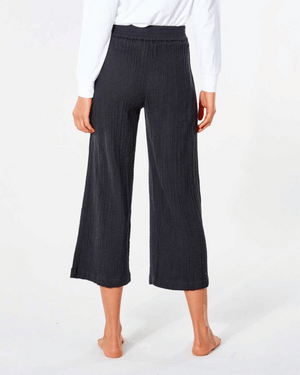 Facing away on a white background, a person is barefoot in black Rip Curl Premium Surf Beach Pants, featuring wide legs and a smocked waist, paired with a white long-sleeve shirt. The cropped pants are ribbed and end at the ankles.