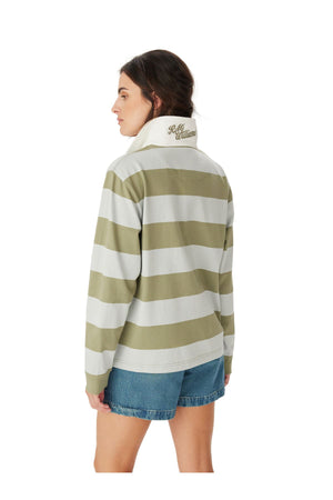 A woman with long brown hair wears the RM Williams Narran Stripe Rugby, a green and white organic cotton shirt with "Hello Williams" on the collar, paired with denim shorts. She stands facing away from the camera.
