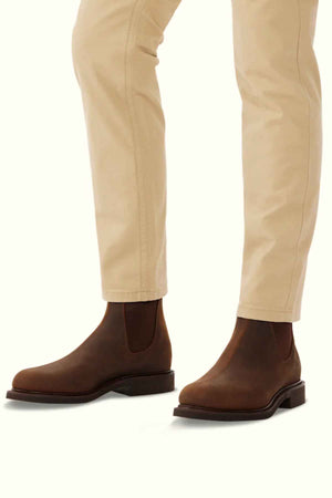 A person in light beige pants and RM Williams Gardener Boots—brown, water-resistant leather Chelsea boots with elastic side panels and a low heel—stands against a plain, light background.