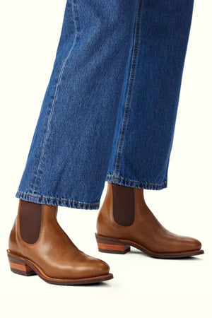 A person in blue, wide-legged jeans wears RM Williams Comfort Lady Yearling Rubber Sole Boots. These brown ankle boots with stacked wooden heels and comfort insoles are shown from the side on a light backdrop, highlighting their stylish design and easy walkability.