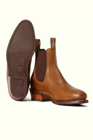 The RM Williams Comfort Lady Yearling Rubber Sole Boot, a women's brown leather Chelsea boot made in Australia, is displayed. With elastic side panels, pull tabs, and a wooden heel, one boot stands upright while the other shows its textured sole for easy wear and timeless style.