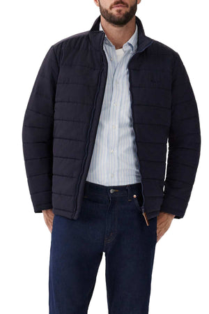 A man wearing the RM Williams Patterson Creek Jacket over a light blue striped shirt and dark blue jeans stands with his hands in his pockets against a white background.