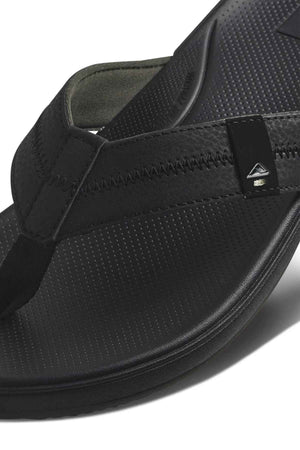 Close-up of a black REEF Cushion Phantom 2.0 flip-flop featuring a textured footbed, visible strap stitching, arch support, and a small triangular metallic logo detail on the side.