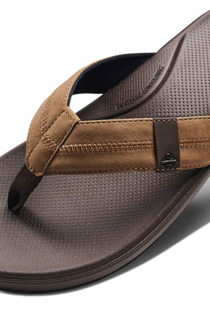 Close-up of a brown REEF Cushion Phantom 2.0 flip-flop with a textured footbed, fabric strap featuring a small metallic logo, detailed stitching, and built-in arch support.