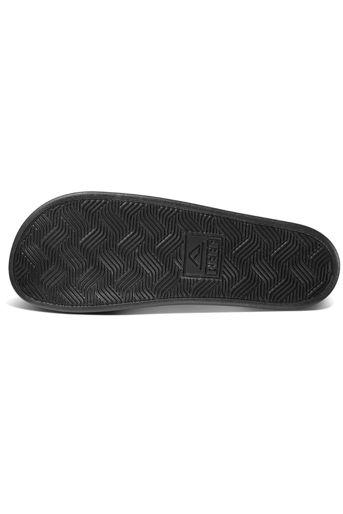 The image displays the black rubber sole of a REEF Cushion Tradewind sandal, featuring a zigzag tread pattern and a rectangular center branding, designed for durable grip similar to classic cushion footbed styles.