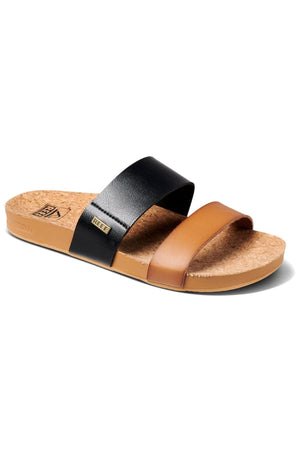 The REEF Cushion Vista Duo sandal features a cork sole and two angled straps, one black and one brown, on a white background. The black strap has a small metal tag with text, and its ergonomic footbed provides cushion bounce comfort for all-day wear.