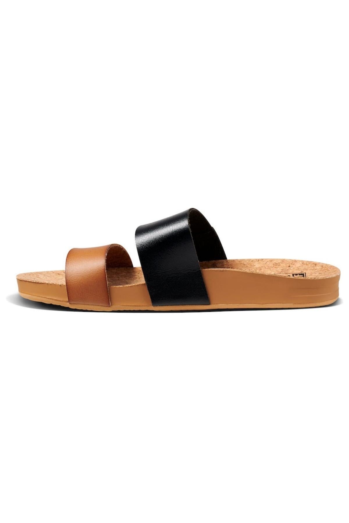 Introducing the REEF Cushion Vista Duo, a slide sandal with a cozy cushion bounce footbed. It has brown and black colors, two wide straps, and is set on a cork-textured brown sole with a durable rubber base. Perfect for all-day wear, combining simplicity and style.