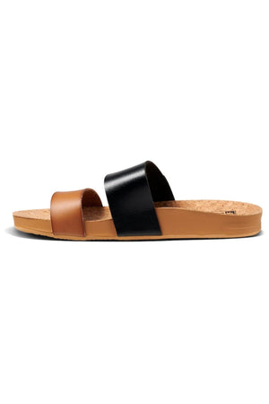 Introducing the REEF Cushion Vista Duo, a slide sandal with a cozy cushion bounce footbed. It has brown and black colors, two wide straps, and is set on a cork-textured brown sole with a durable rubber base. Perfect for all-day wear, combining simplicity and style.