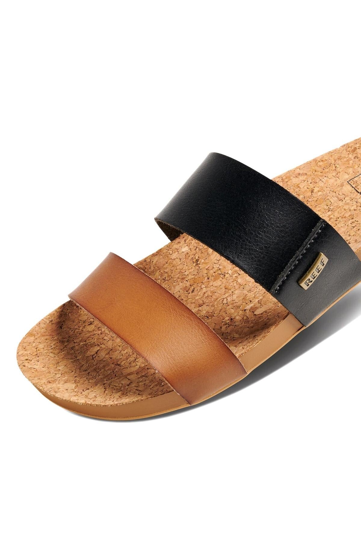 A close-up of the REEF Cushion Vista Duo sandal features black and brown wide straps across the foot, a visible label on the black strap, a tan footbed, and a cork sole for cushion bounce comfort.
