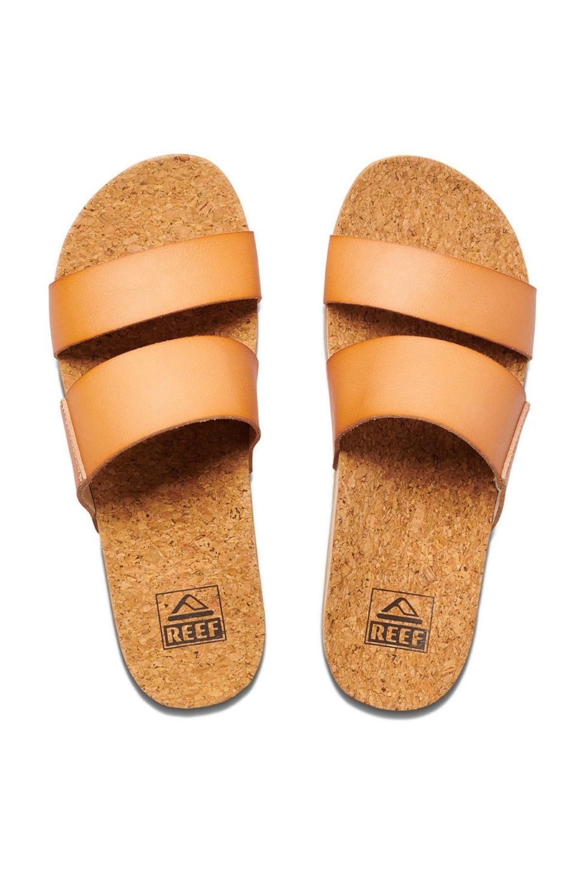 A pair of tan REEF Cushion Vista Hi slip-on sandals with two wide straps across the top, featuring cork insoles and a subtle platform heel. The brand name "REEF" and its logo are visible on the cushioned insole.