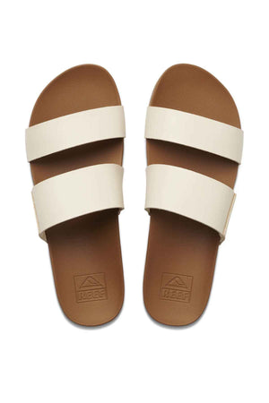A pair of REEF Cushion Vista brown slide sandals with minimalist design features two wide cream straps, shown from above. The footbed offers memory-free foam and arch support, with the brand name embossed on it.