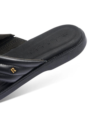 Close-up of the REEF Lofty Lux X sandal, showing a padded strap with a gold "R" detail, visible stitching, and textured material. This sustainable sandal offers premium comfort and features embossed text on the footbed.