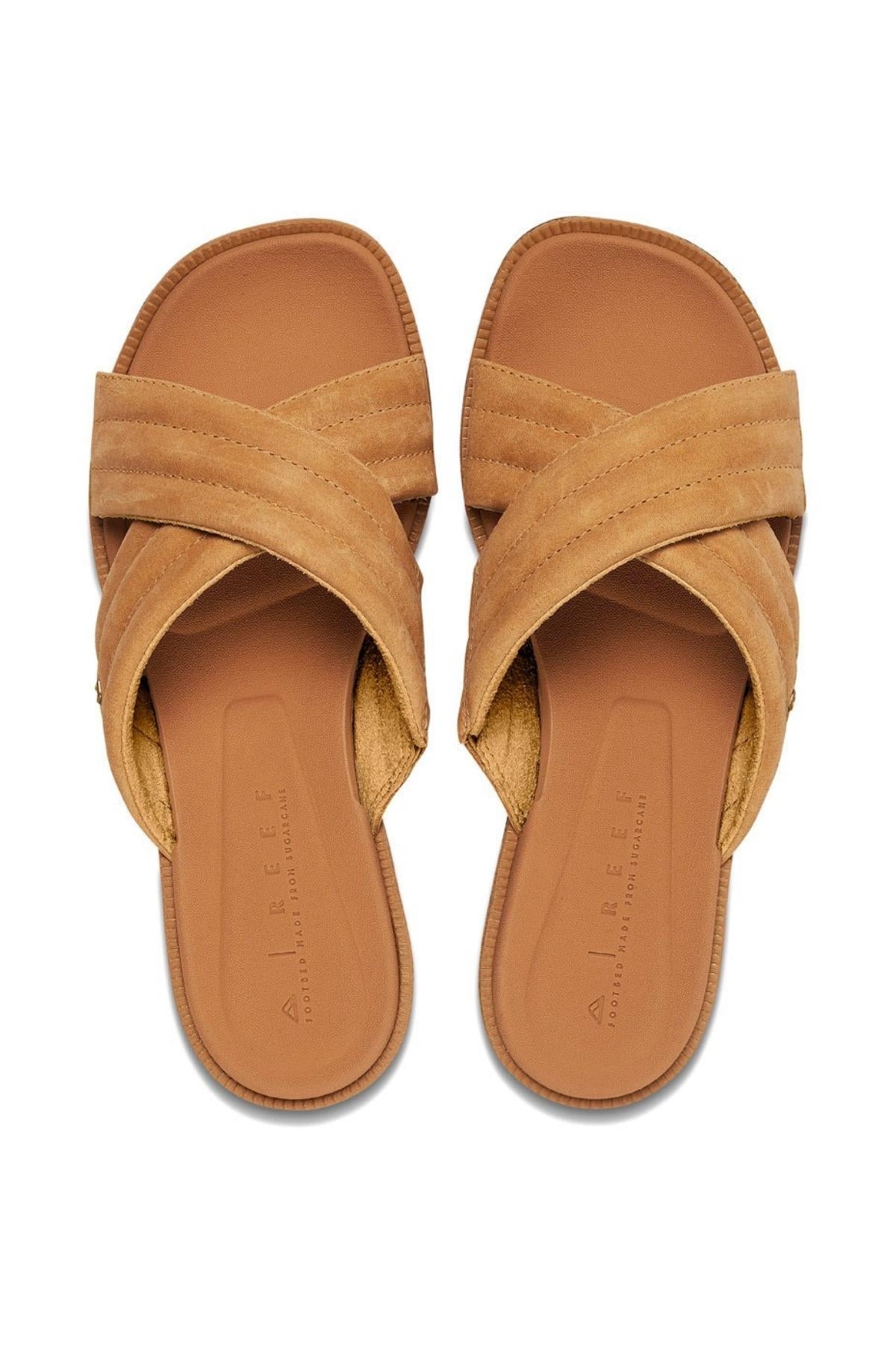REEF Lofty Lux X slide sandals feature crisscross straps on a light brown footbed for premium comfort. The visible brand text highlights their casual appeal and sustainable design, showcased side by side.