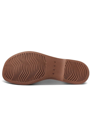 The image displays the sole of a REEF Lofty Lux X shoe, showing a textured pattern with wavy grooves for grip. This brown sole represents premium comfort and sustainability, with "REEF" embossed near the arch.