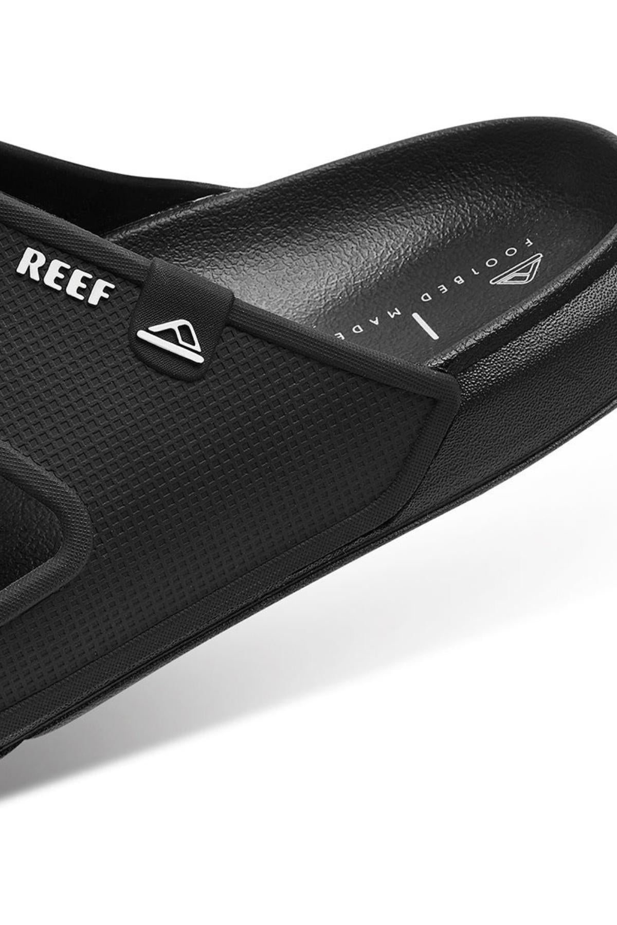 Close-up of REEF Oasis Double Up black slide sandal with textured material, featuring the "REEF" brand name and a triangle logo. The water-friendly sandal shows partially visible "FOOTBED MADE," indicating its comfortable arch support.