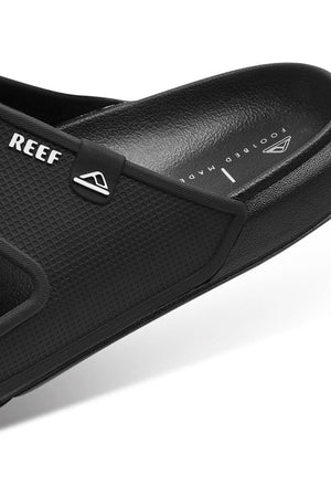 Close-up of REEF Oasis Double Up black slide sandal with textured material, featuring the "REEF" brand name and a triangle logo. The water-friendly sandal shows partially visible "FOOTBED MADE," indicating its comfortable arch support.