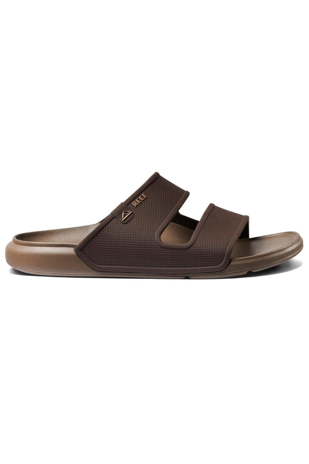 The REEF Oasis Double Up is a brown slide sandal featuring two wide straps, a textured footbed made from renewable sugarcane EVA for arch support, and the Reef logo on the upper strap.