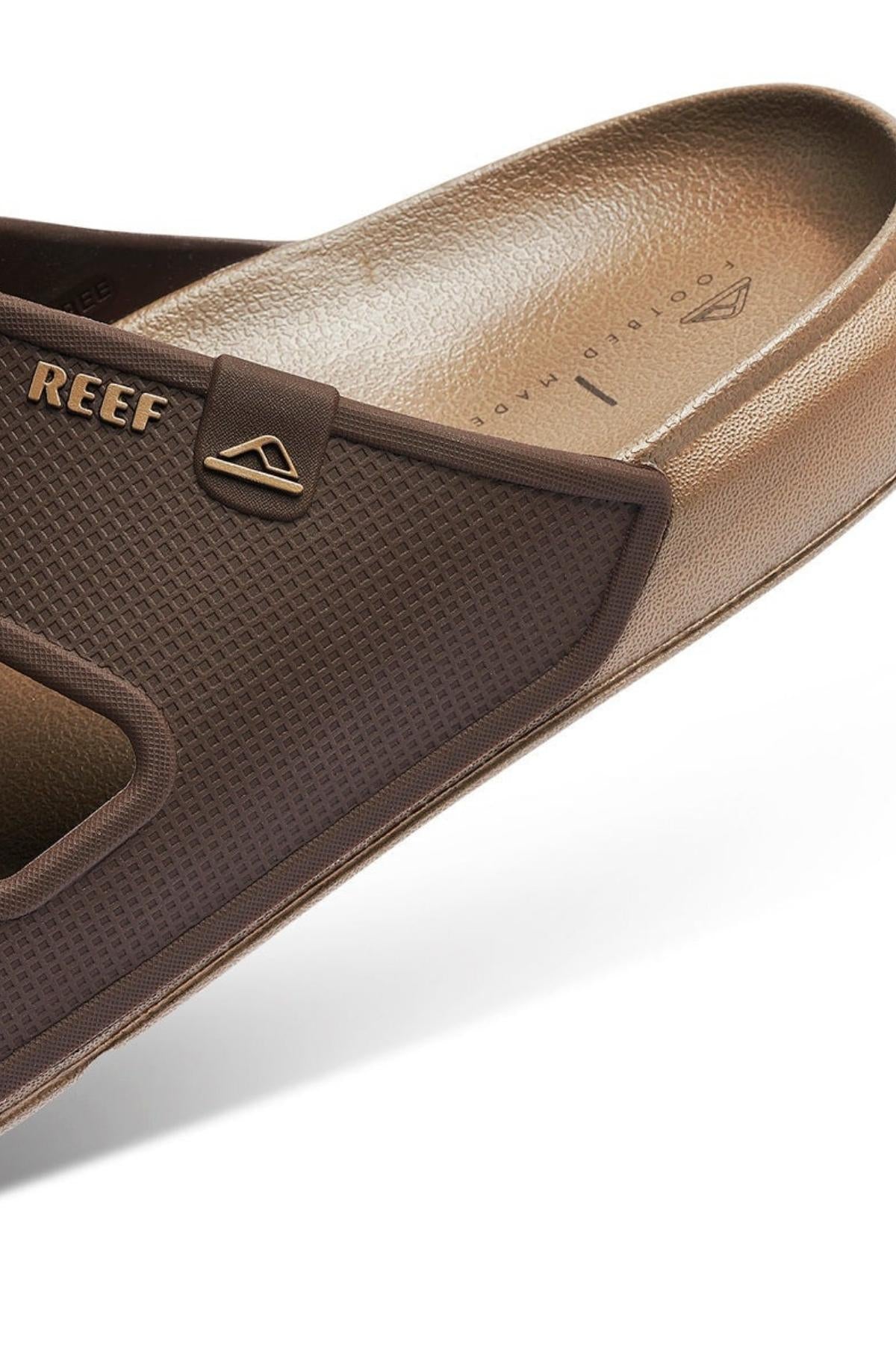 Close-up of the REEF Oasis Double Up sandal in brown, featuring textured material, brand logo, and a renewable sugarcane EVA footbed with arch support on a white background.