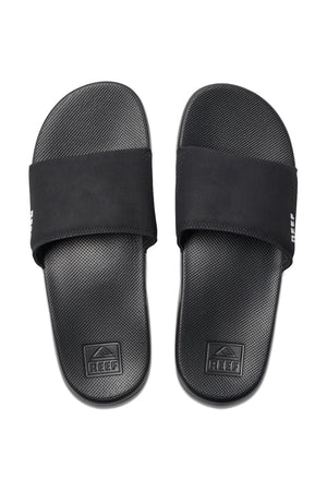 The REEF One Slide sandals are black, slip-on with a wide strap over the top. Featuring a textured footbed for added comfort, these stylish slides prominently display the "REEF" branding on the outer side of each strap and on the heel area of the footbed, emphasizing their waterproof design for versatile use.