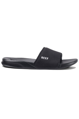 The REEF One Slide features a sleek, black design with a contoured footbed and wide strap across the top of the foot. The strap has small perforations for ventilation and proudly displays the white "REEF" logo. These stylish slides offer modern comfort with a waterproof design, perfect for versatile wear.