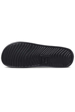 The image showcases the underside of a REEF One Slide in black, featuring a wavy tread pattern characteristic of REEF shoes. In the central region near the heel, there's a rectangular section, likely indicating a logo or brand name. The sole's design seems to prioritize traction and grip, highlighting its waterproof properties.