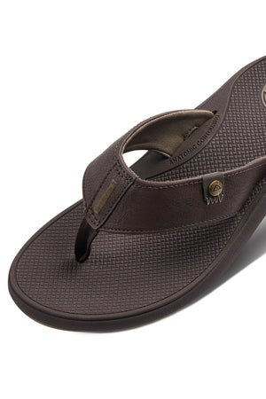 Close-up of the REEF Phantom Nias flip-flop sandal in brown, showcasing its textured footbed and synthetic upper with brand logos and stitching details. This sandal features superior traction capabilities, an ergonomic design, and a toe thong strap for added comfort.