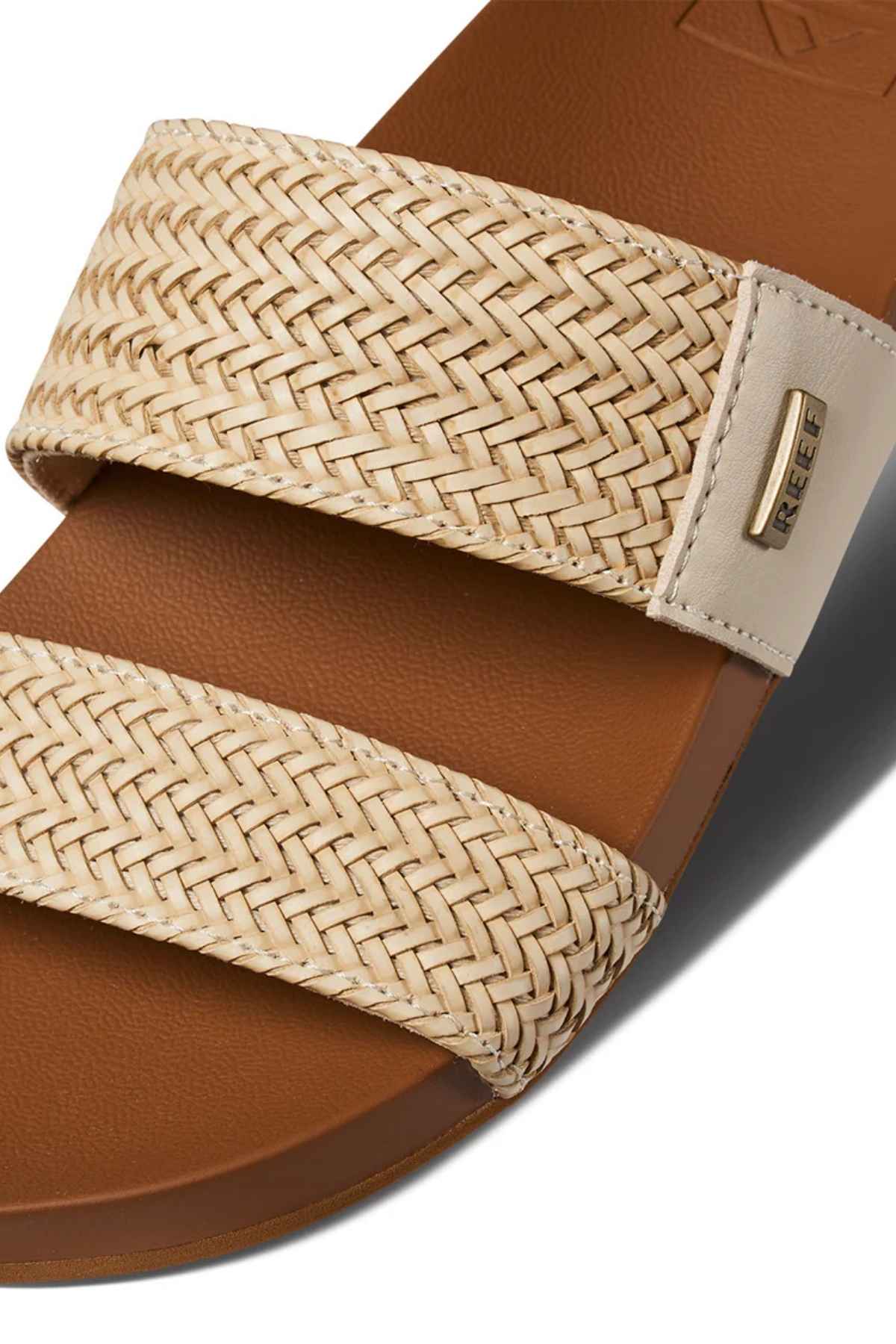 Close-up of the REEF Vista Braid II sandal, showcasing woven beige straps, a brown sole, a small metal "REEF" tag on the top strap, and a cushioned midsole for extra comfort.