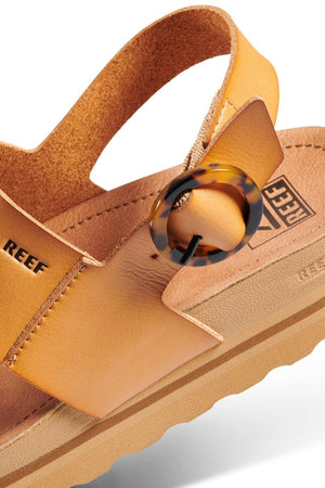 Close-up of a tan sandal with a brown tortoiseshell-style circular buckle. The REEF Vista Hi Buckle sandals feature a vegan leather upper, cushioned sole, and textured insole with thoughtfully integrated arch support. The brand name "REEF" is visible on the strap and insole, highlighting its modern design.