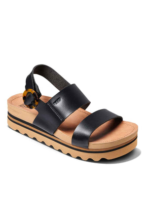 The REEF Vista Hi Buckle is a black vegan leather sandal featuring two wide straps, an adjustable heel buckle, a tan cushioned sole, and a wavy black outsole.