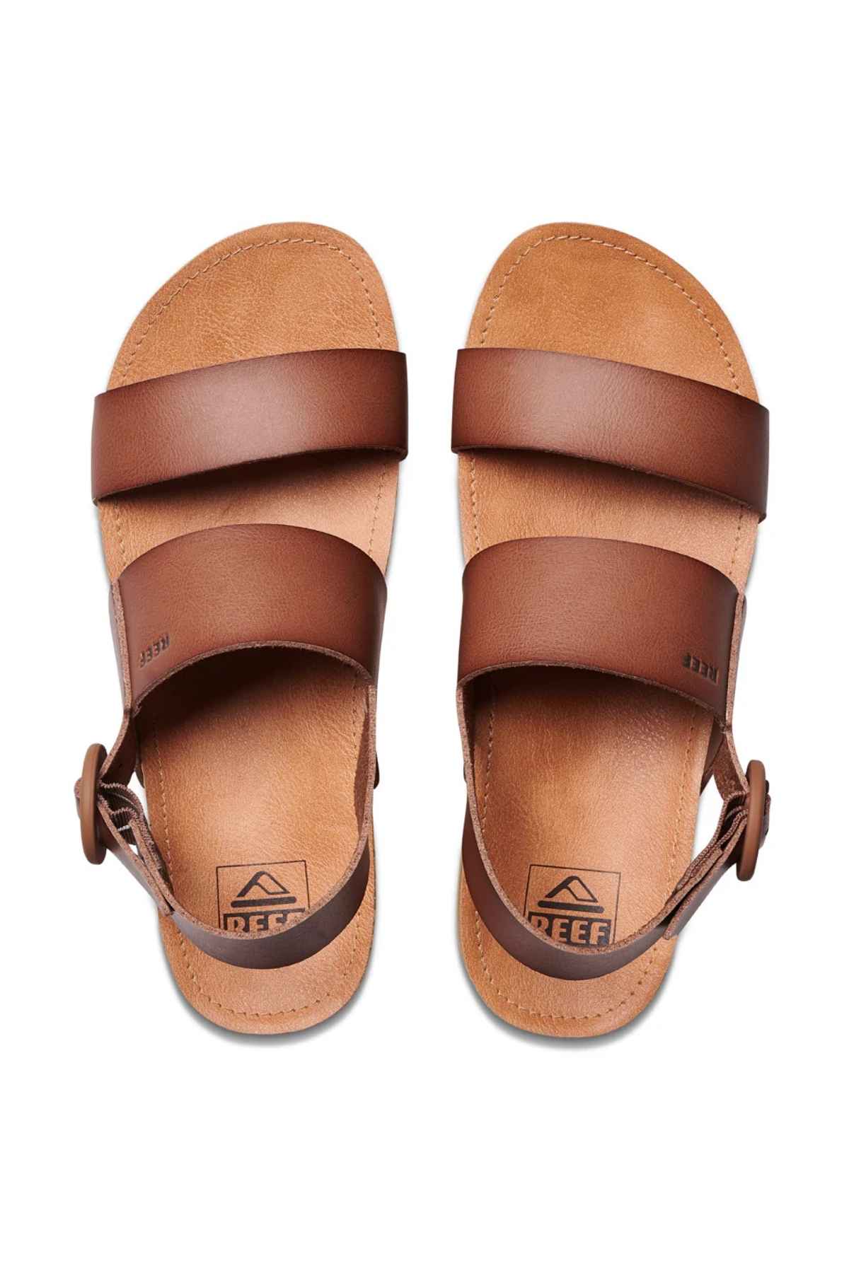 The REEF Vista Hi Buckle sandals showcase brown vegan leather straps, with two crossing over the foot and an ankle strap featuring a buckle. The soles are adorned with an embossed logo. Photographed from a top-down angle against a plain background, these platform sandals combine style and sustainability.