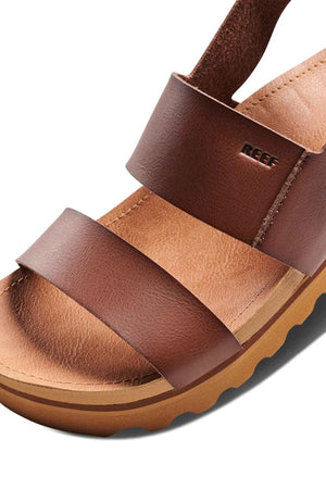 Close-up of the REEF Vista Hi Buckle sandal in brown, featuring two wide vegan leather straps embossed with "REEF" and a tan cushioned sole with a slightly elevated, curvy design.