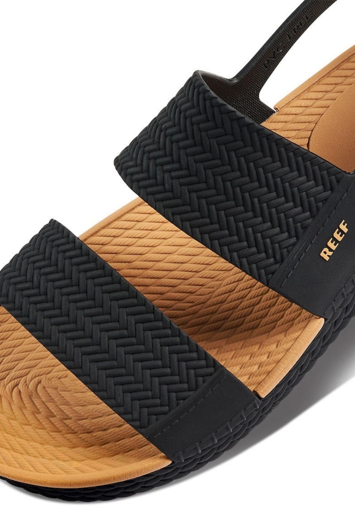 A close-up of the REEF Water Vista sandal showcases black woven straps and a tan footbed with arch support. The textured sole provides grip, while "REEF" gleams in gold on the side, embodying the brand's style and comfort.