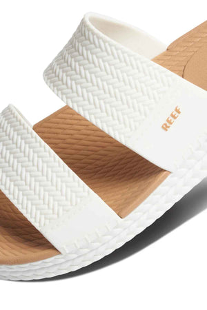 Close-up of a REEF Water Vista Slide sandal in white with a woven texture, two thick straps, gold "REEF" branding, and a light brown dual-density EVA footbed featuring a textured pattern for comfort.