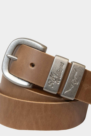 The RM Williams Drover Belt is a rolled-up brown solid hide leather belt with a brushed nickel buckle, engraved branding, script text, and a flying eagle design on the metal loop; proudly made in Australia.
