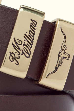 Close-up of the RM Williams 1 1/2 3 Piece Solid Hide Belt, crafted from brown leather and adorned with gold hardware. This premium hide leather belt features a beautifully engraved brass buckle, showcasing the signature "R.M. Williams" on one side and an image of a longhorn skull on the other. Made in Australia, this piece exemplifies exceptional craftsmanship.