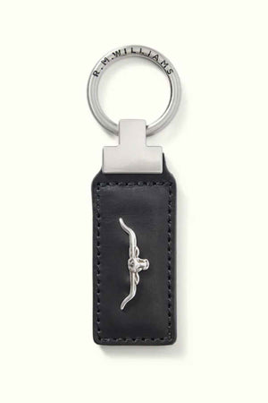 The RM Williams Angaston Key Fob is crafted from black full-grain leather with intricate stitching and showcases a silver metal longhorn emblem at its center. The silver keyring loop is engraved with "R.M. WILLIAMS.