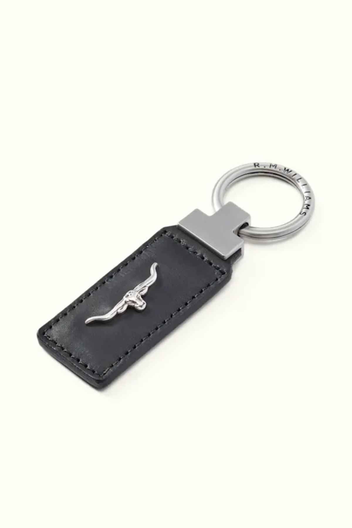 The RM Williams Angaston Key Fob features a silver ring engraved with "R.M.W." and is attached to a black rectangular fob made from full-grain leather, embellished with a metal bull emblem.