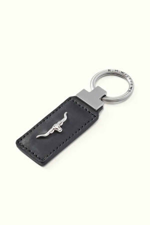 The RM Williams Angaston Key Fob features a silver ring engraved with "R.M.W." and is attached to a black rectangular fob made from full-grain leather, embellished with a metal bull emblem.