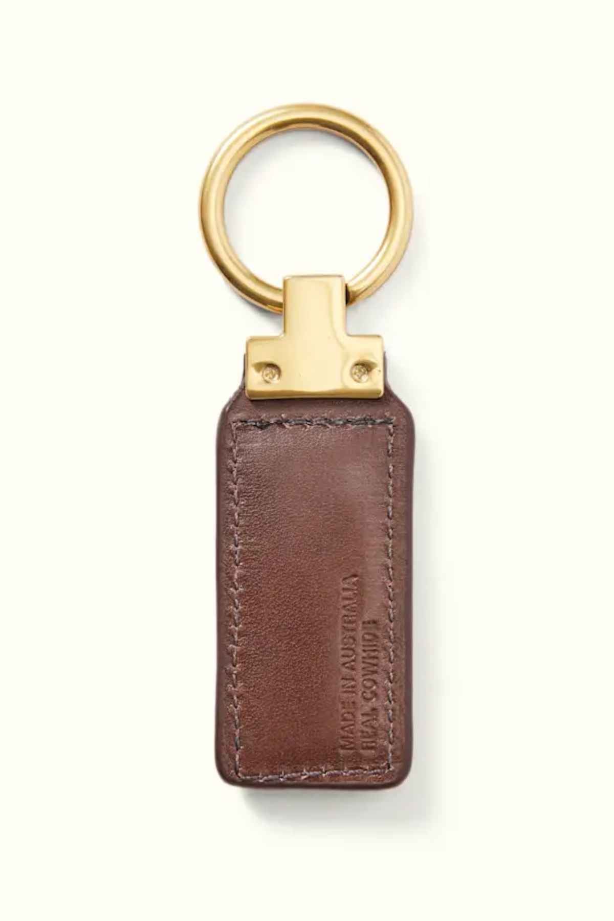 The RM Williams Angaston Key Fob features a brown leather design with an antique brass ring and top. The full-grain leather is embossed with text, and visible stitching along the edges highlights its quality craftsmanship.