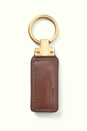 The RM Williams Angaston Key Fob features a brown leather design with an antique brass ring and top. The full-grain leather is embossed with text, and visible stitching along the edges highlights its quality craftsmanship.