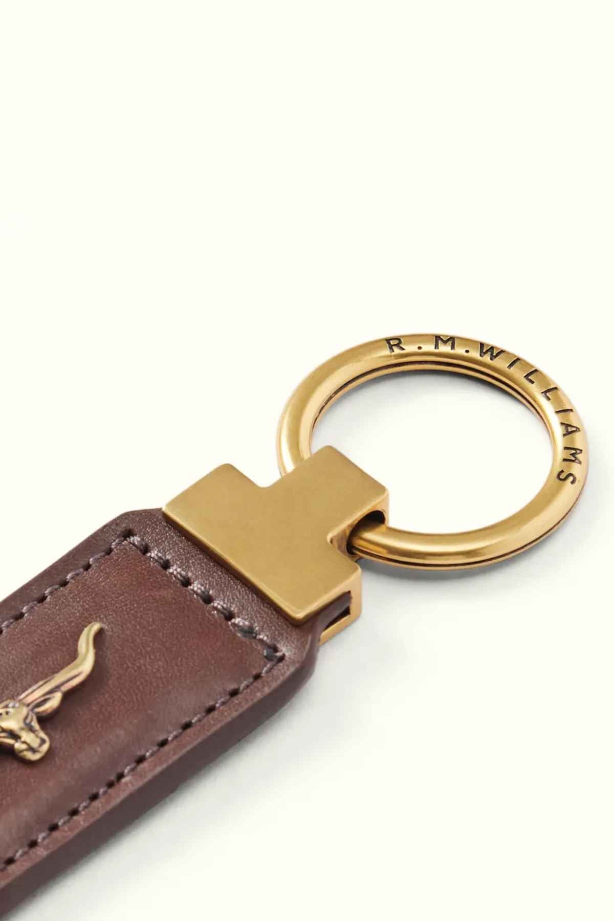 A detailed image of the RM Williams Angaston Key Fob, crafted from brown, full-grain leather and adorned with an antique brass embellishment. The engraved metal ring reads "R.M. Williams" and boasts a polished finish. This Australian-made piece is connected to the leather by a rectangular gold connector.